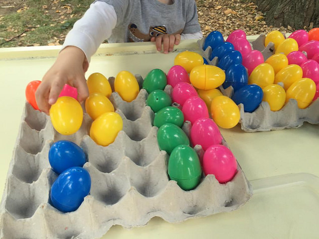 Small child putting colorful plastic eggs into a bulk egg carton in a semi-organized fashion.