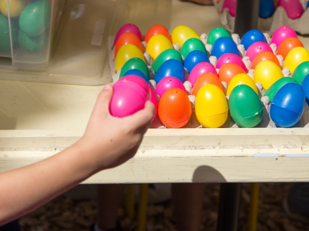 A childs hand places a pink egg in a carton of colorful plastic eggs.
