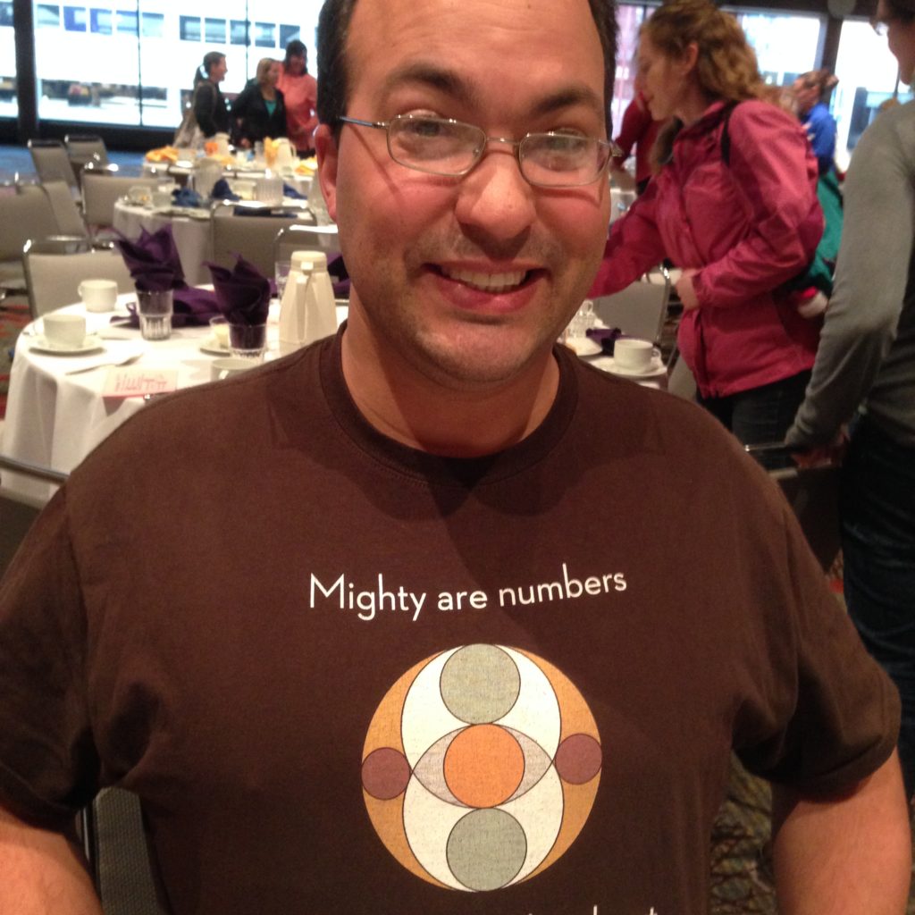 Jonathan Osters modeling a shirt that says "Mighty are numbers" and featuring a design of circles overlapping each other within a larger circle