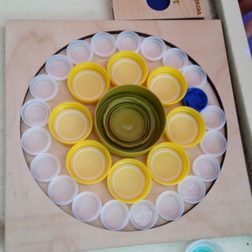 One large green bottle cap surrounded by a bunch of medium yellow ones, and that whole thing surrounded by a lot of small white ones and one small blue bottle cap—all inside a large circular frame