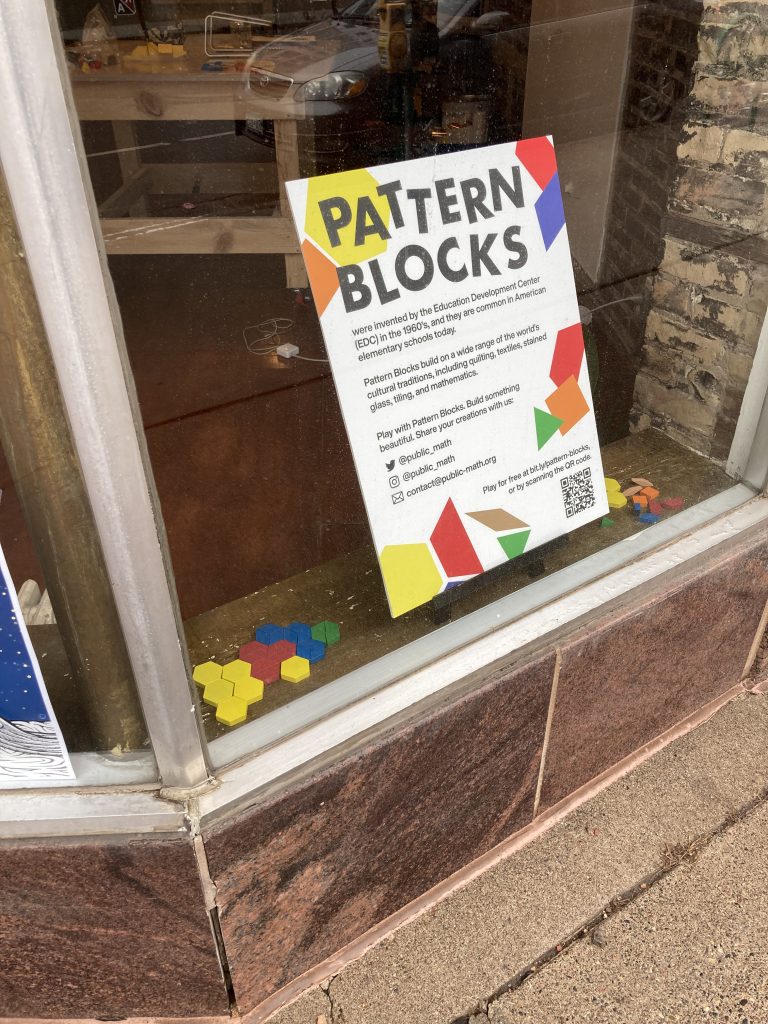 Close up view of the smaller window. The sign reads "Pattern Blocks were invented by the Education Development Center (EDC) in the 1960’s, and they are common in American elementary schools today. Pattern Blocks build on a wide range of the world’s cultural traditions, including quilting, textiles, stained glass, tiling, and mathematics. Play with Pattern Blocks. Building something beautiful. Share your creations with us: @public_math on Twitter and Instagram, or by email: contact@public-math.org Play for free at bit.ly/pattern-blocks, or by scanning the QR code."