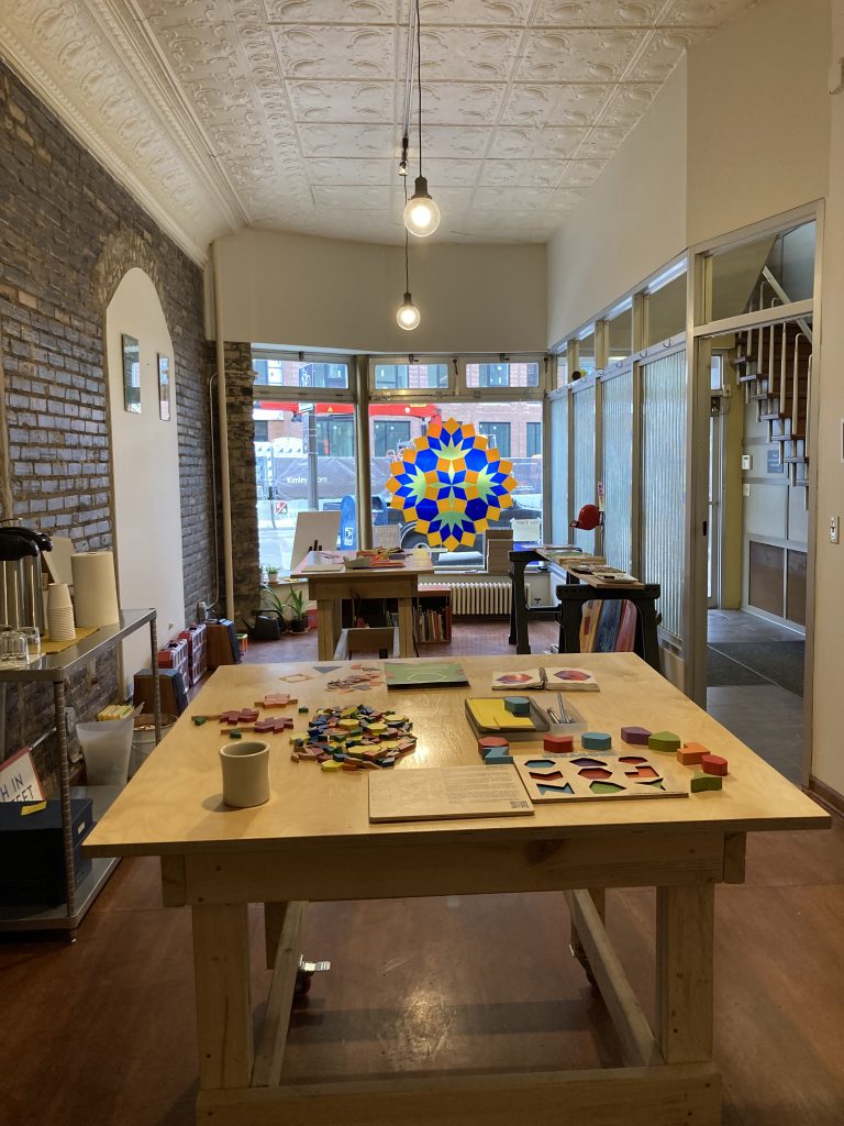 Interior view of a long, thin room. The windows we saw earlier are at the far end, and there are tables with colorful math toys arranged invitingly. One wall is exposed brick, and there is a high tin ceiling.