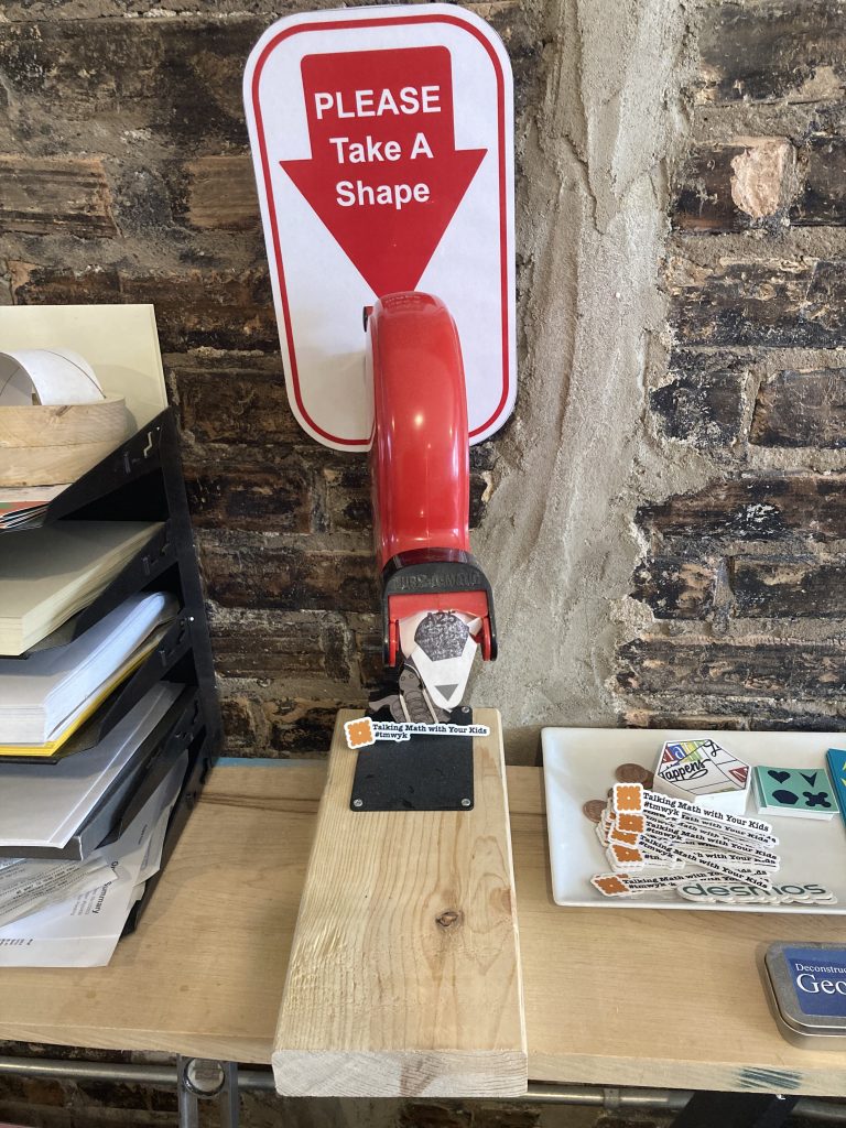 A "Please Take a Number" machine as you might see at a deli counter, but this one says "Please Take a Shape". A ticket with a sort of muffin shape in place of a number sticks out the front of the machine.