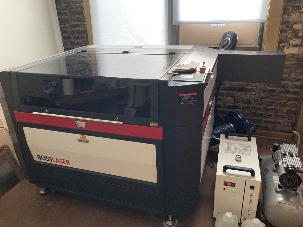 A large black, red, and white laser cutter (and associated machinery) in front of two tall windows.