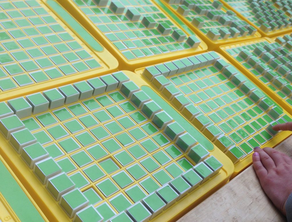 A child plays with a grid of machines, each containing a grid of green square buttons that pop up and down. The child is making a large square on one of the machines.