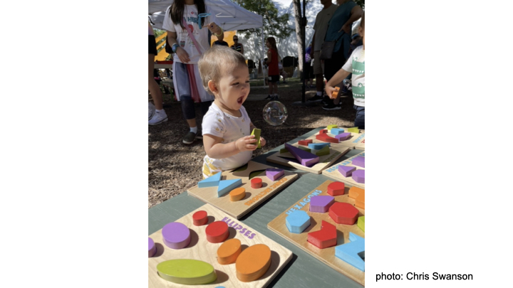 A very happy toddler playing with colorful shapes puzzles outside on a sunny day. A large bubble floats right in front of her. photo credit: Chris Swanson