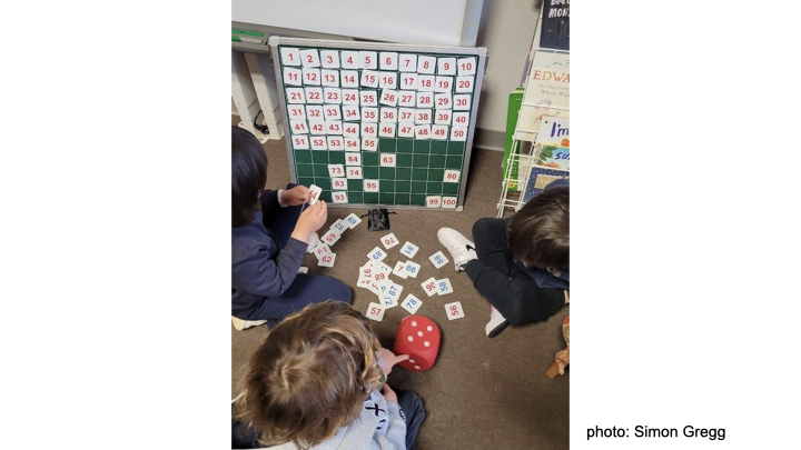 Three young children play with a gridded board and magnetic squares. Each square has a number. They seem to be arranging the squares in 10 rows of 10, and have completed the first five and a half rows, with a number of scattered larger numbers in place on the bottom half of the grid.