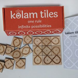 A brightly colored, closed reading "kolam tiles: one rule, infinite possibilities" sits on a table.Several of laser-engraved, square wooden tiles tiles are scattered haphazardly to the left.. On the left there is a three-by-three tile tile arrangement that results in several intertwined and overlapping paths. There is a sheet of paper that reads "kolam tiles; a tiny guide"