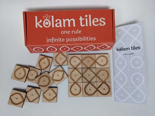 A brightly colored, closed reading "kolam tiles: one rule, infinite possibilities" sits on a table.Several of laser-engraved, square wooden tiles tiles are scattered haphazardly to the left.. On the left there is a three-by-three tile tile arrangement that results in several intertwined and overlapping paths. There is a sheet of paper that reads "kolam tiles; a tiny guide"