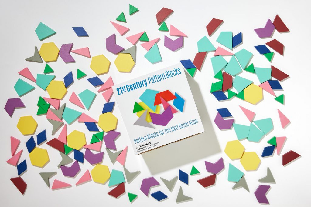 A large collection of colorful polygons, scattered on a white surface and brightly lit Looking almost like candy, there are pink right triangles, yellow hexagons, purple irregular hexagons, blue diamonds, red isosceles trapezoids, gray darts, and (best of all) teal kites. In the center of the image is the box these tiles come in, which reads "21st Century Pattern Blocks. Pattern Blocks for the Next Generation"