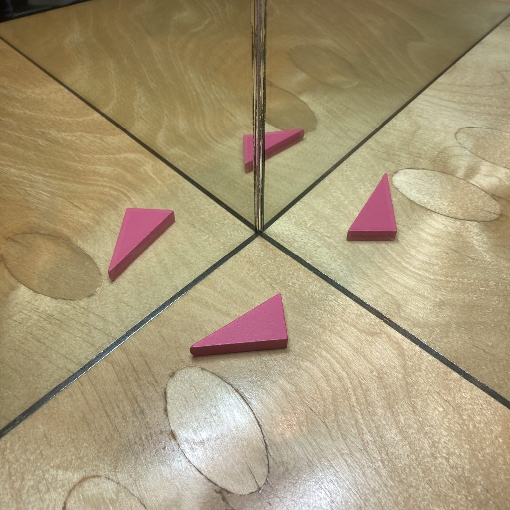 one pink right triangle, lying on a polished wooden table, pointing to the left. There are two hinged mirrors, opened to a right angle, and reflecting the triangle in each. There is a fourth triangle in the reflections of the reflections