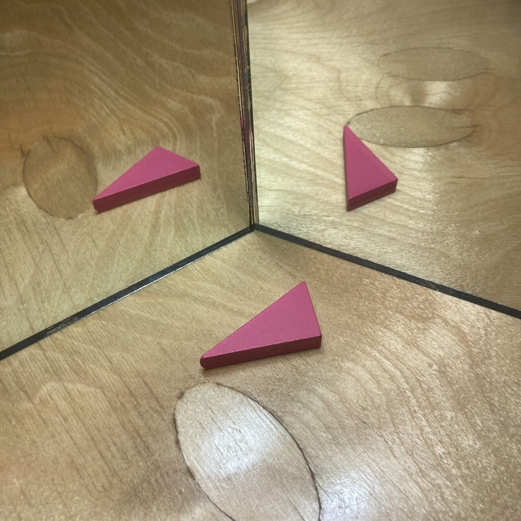 one pink right triangle, lying on a polished wooden table, pointing to the left. There are two hinged mirrors, opened to an obtuse angle, and reflecting the triangle in each