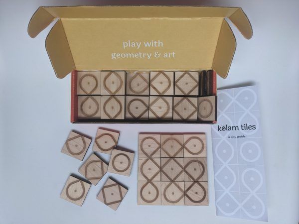 An open box of laser-engraved square wooden tiles. Several of the tiles are scattered haphazardly to the left.. On the left there is a three-by-three tile tile arrangement that results in several intertwined and overlapping paths. There is a sheet of paper that reads "kolam tiles; a tiny guide"