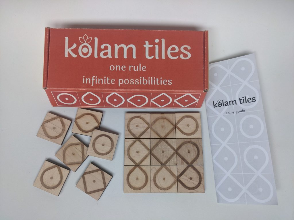 A brightly colored, closed box reading "kolam tiles: one rule, infinite possibilities" sits on a table.Several of laser-engraved, square wooden tiles tiles are scattered haphazardly to the left.. On the left there is a three-by-three tile tile arrangement that results in several intertwined and overlapping paths. There is a sheet of paper that reads "kolam tiles; a tiny guide"