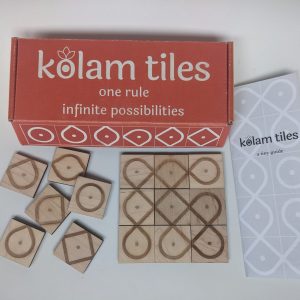 A brightly colored, closed box reading "kolam tiles: one rule, infinite possibilities" sits on a table.Several of laser-engraved, square wooden tiles tiles are scattered haphazardly to the left.. On the left there is a three-by-three tile tile arrangement that results in several intertwined and overlapping paths. There is a sheet of paper that reads "kolam tiles; a tiny guide"