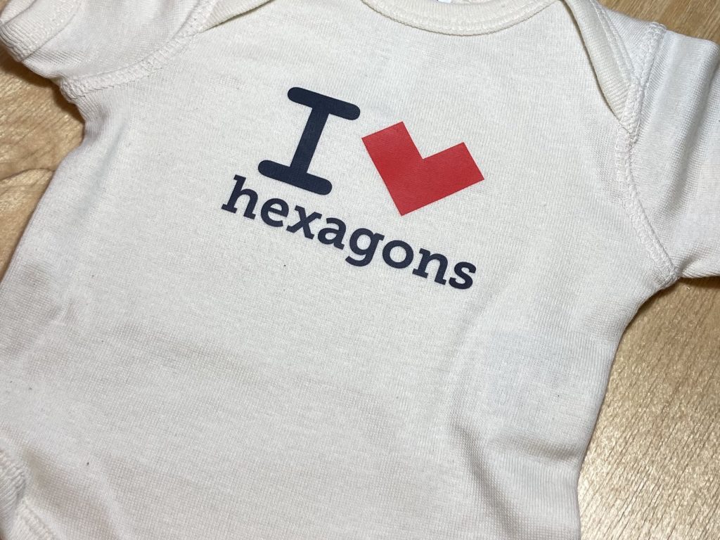An off-white baby bodysuit that reads "I *heart* hexagons" but where the heart is actually a red, concave hexagon standing on a vertex.