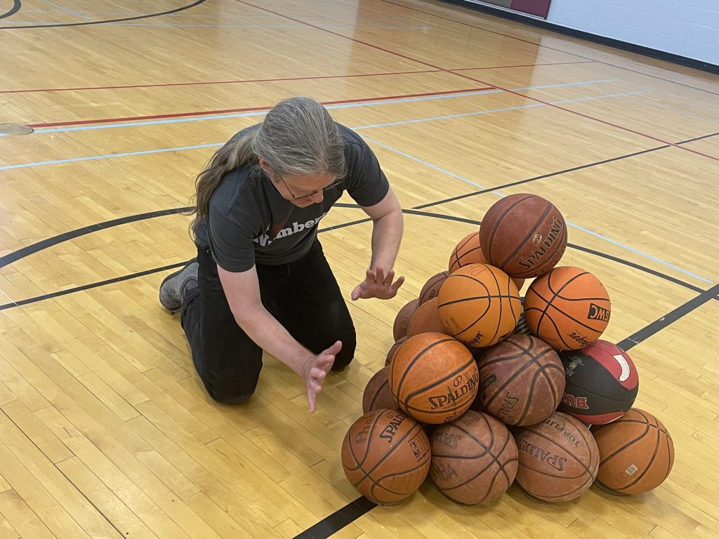 I am kneeling on the wooden floor of a gymnasium, holding my hands as if to encourage the tetrahedron of basketballs in front of to behave and not roll away. The tetrahedron is four layers of basketballs tall.