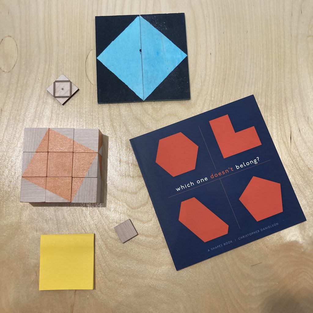 A photograph of a number of square objects on a tabletop. There is a square made of two rectangular tiles, which also combine two painted right triangles to make an interior square. There is a Post-It, and a square Which One Doesn't Belong? book.